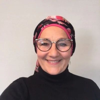 A white woman wearing glasses and a colorful head scarf, smiling softly gazes directly at the viewer, exuding warmth and confidence.